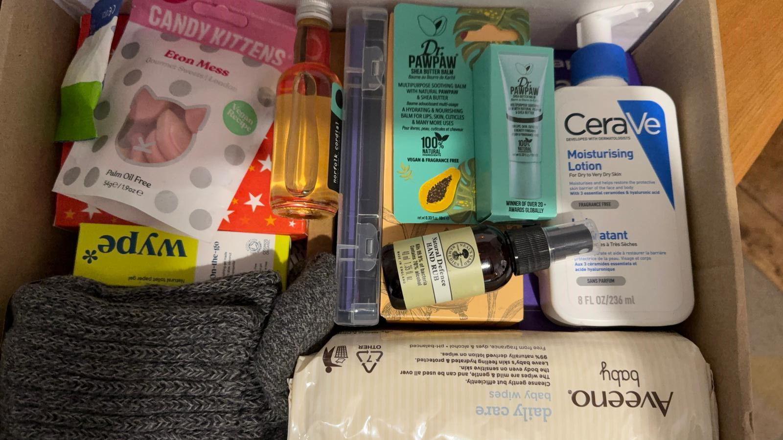 An open cardboard box containing closely-packed items: Aveeno baby wipes, CeraVe Moisturing Lotion in a pump-action white bottle, Candy Kittens Eton Mess sweets in a resealable pink and white pouch, grey woollen socks, Dr PawPaw balm in a tourquoise box, a small bottle of yellow cordial.