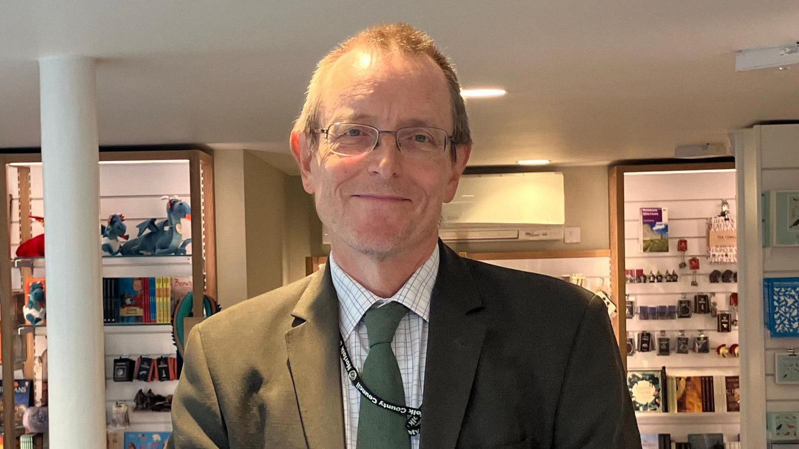 Dr Robin Hanley standing inside a room in Norwich Castle. He is wearing a dark green suit jacket, with a checked shirt and dark green tie. He is wearing glasses and is looking directly at the camera and smiling.