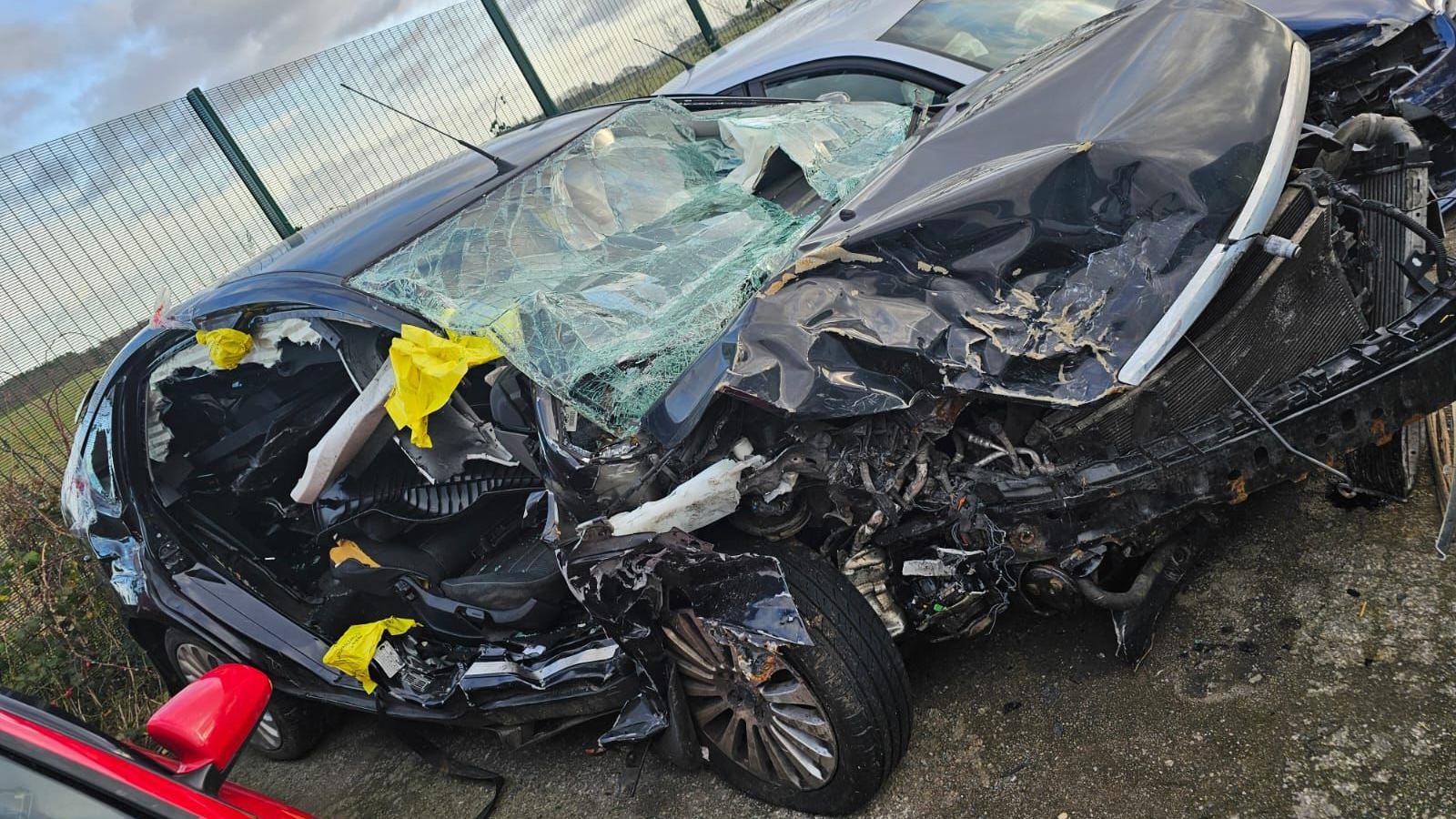 The wreckage of Tony's car. The black bodywork is crumpled and badly damaged. The driver's door and passenger door have been cut away.