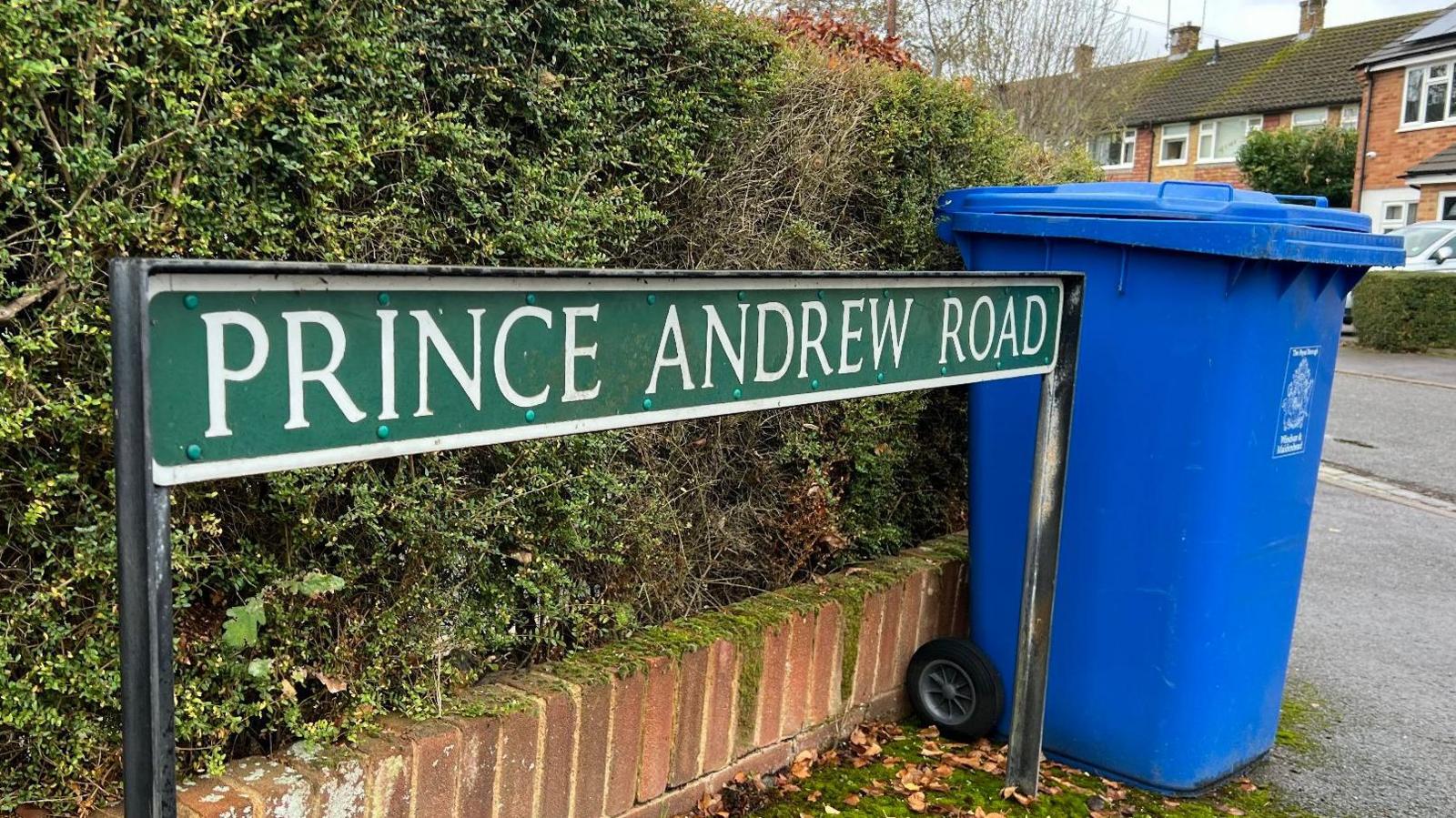 The image shows the road sign for Prince Andrew Road. Next to it is a blue wheelie bin. Behind the sign is a hedge. 