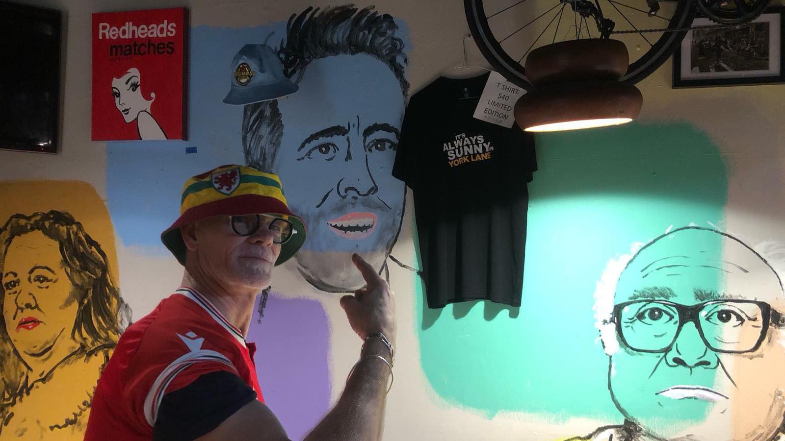 Dieter pointing at the wall of the pub which has drawings connected to Wrexham. He is pointing at Ryan Reynolds, on the right is Danny DeVito in a nod to a hit show Rob McElhenney stars in. There is also a woman on the left of Dieter drawn on the wall.