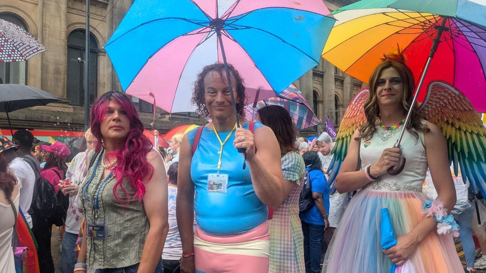 Three people walking down a city street as others mill about in the background. The middle person of the three is wearing blue and pink and is holding an umbrella sporting the same colours. To their left is someone holding a rainbow-coloured umbrella and wearing a white dress and rainbow wings. On the other side is someone with long, flowing pink hair.