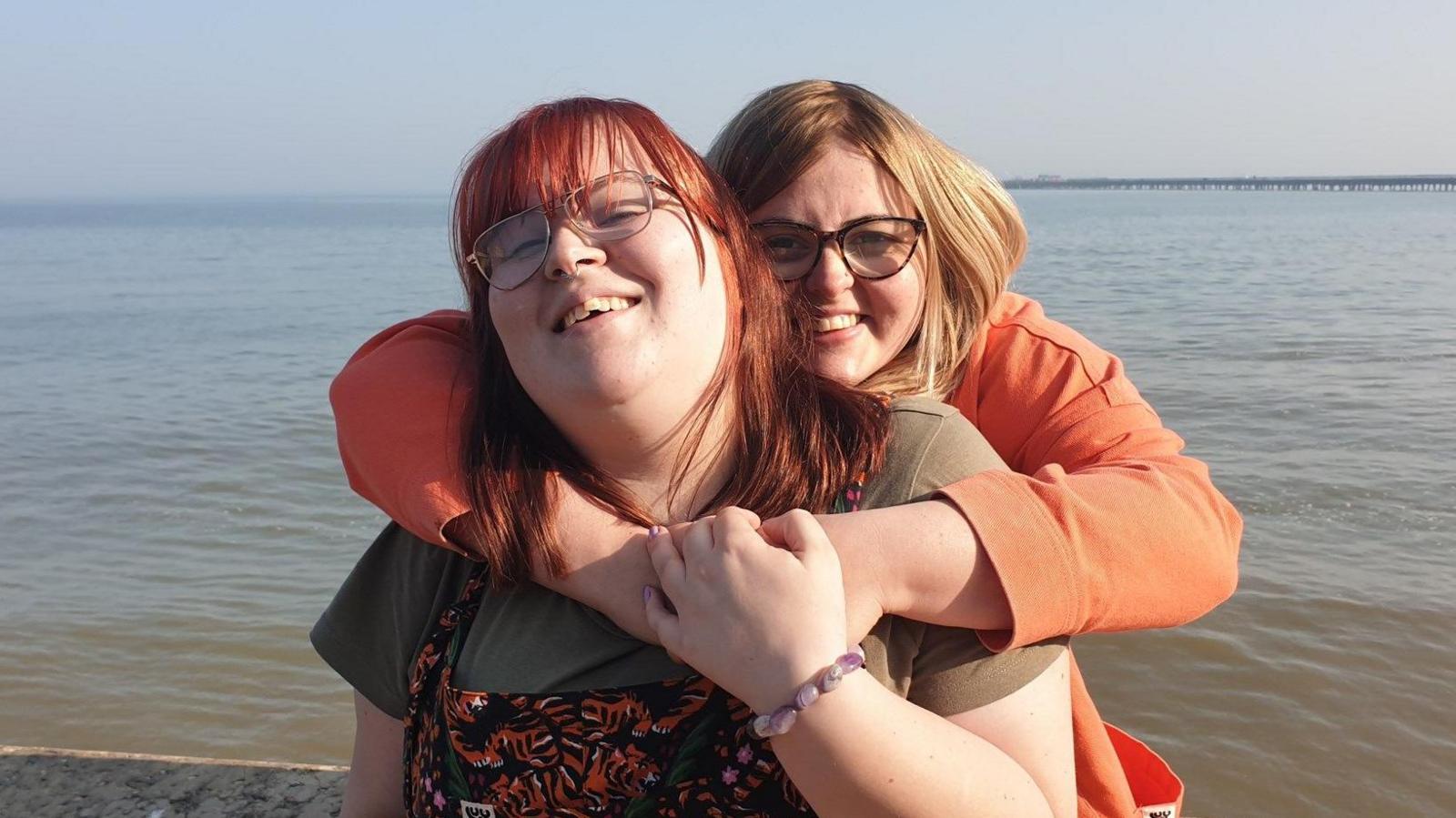 Zoe is being hugged by a woman from behind as they stand in front of a large body of water, possibly the sea. Zoe has long red hair, a full fringe, and is wearing glasses, a green top, and an orange patterned dress. Mrs Nunn has blonde hair, a side fringe, and is wearing an orange coat and glasses. Both women are smiling.