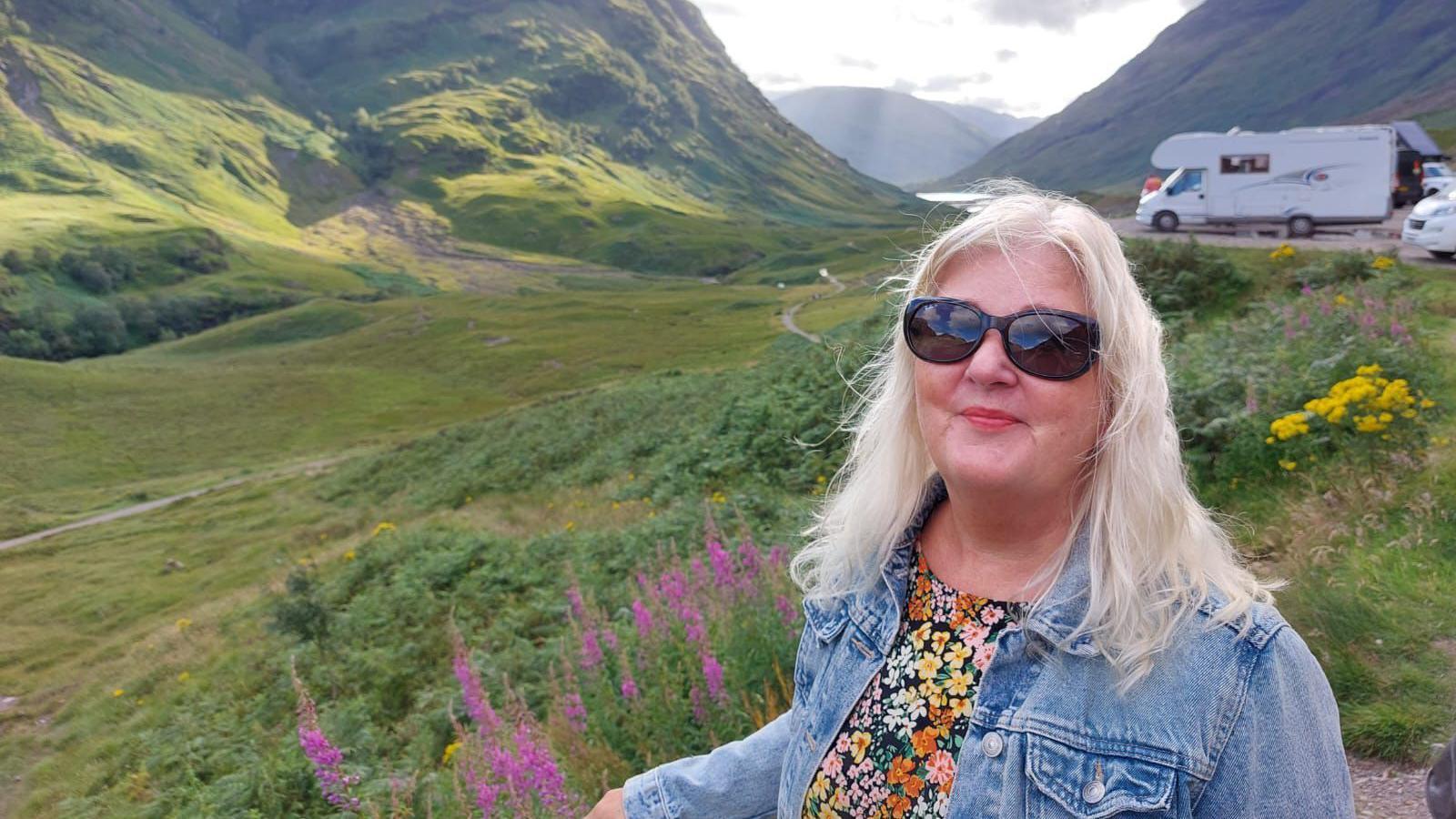 Nicky Maidment has medium length bleach blonde hair and wears a floral top, denim jacket and sunglasses.
She stands in a meadow near some mountains.