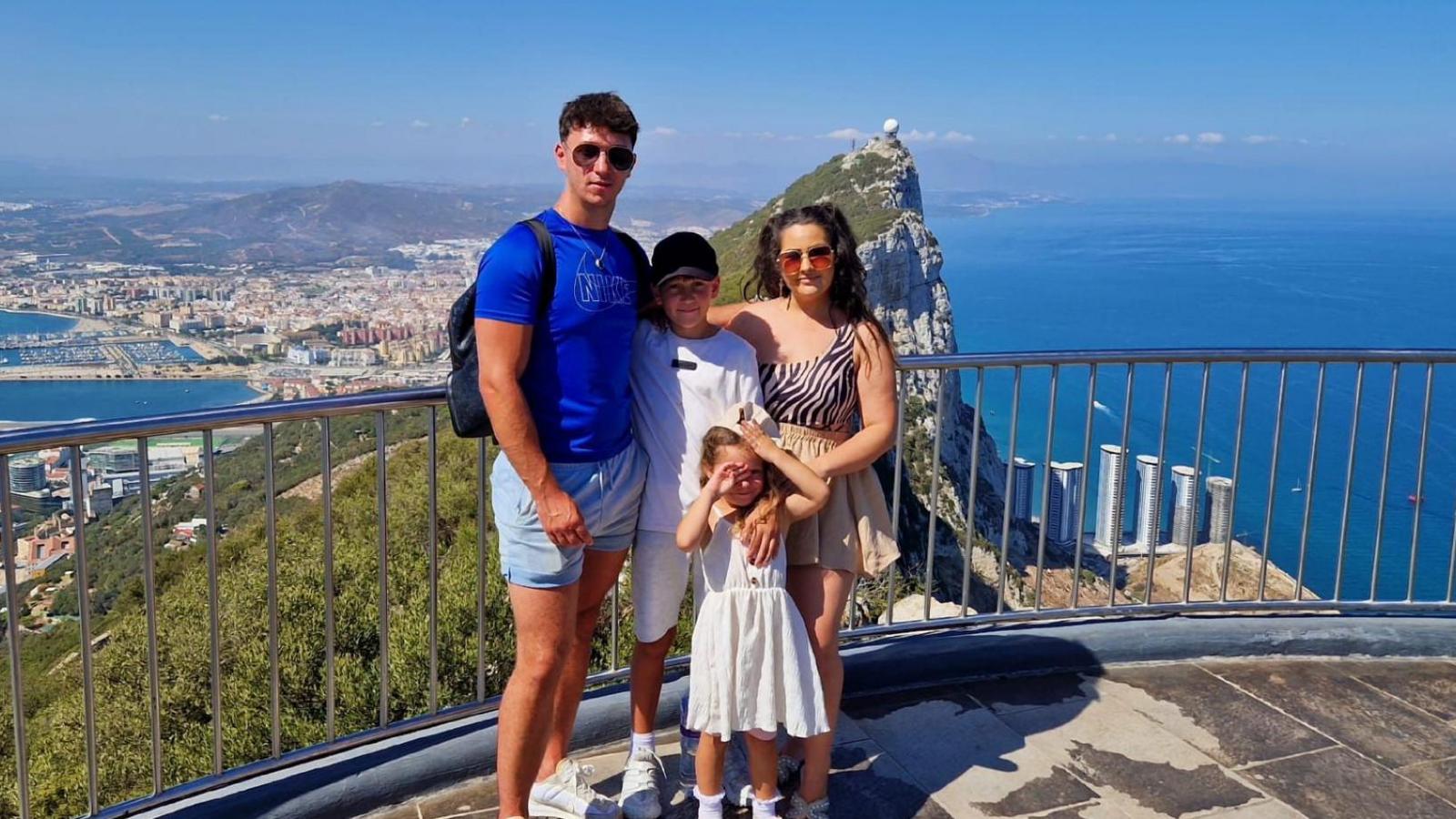 A family of four stand on a platform overlooking a mountain and coastline. On the left is a tall man wearing a blue top and sunglasses, in the middle is a boy with a black cap and white top, on the right is a woman in sunglasses and an animal print top and in front is a young girl in a white dress