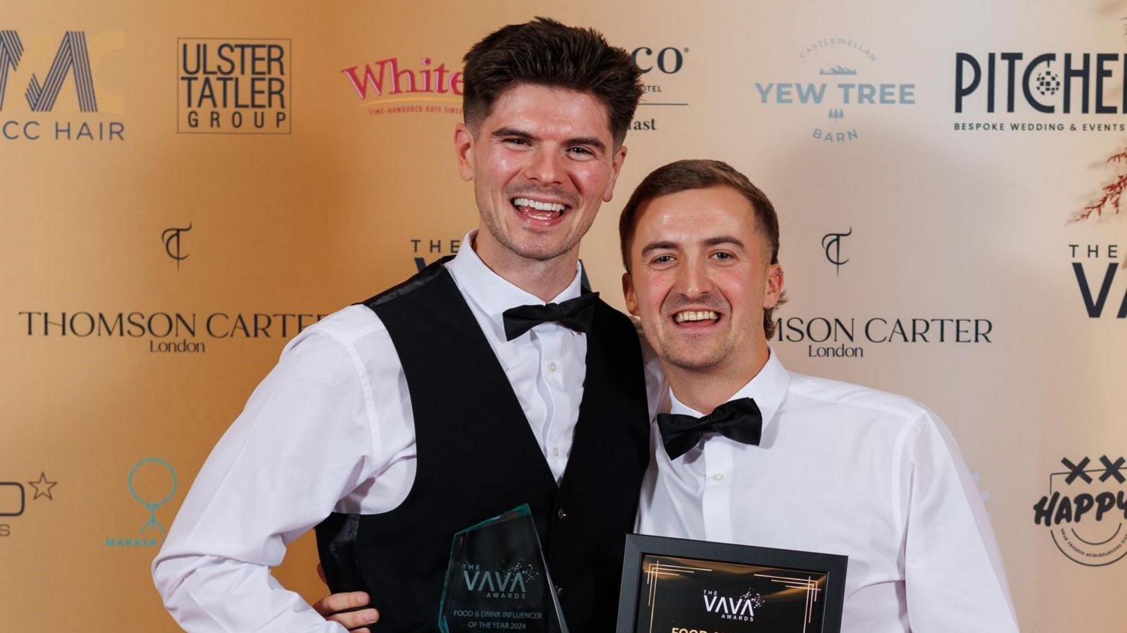 Two men wearing tuxedos smiling at the camera with their arms around each other in front of a background with multiple corporate logos