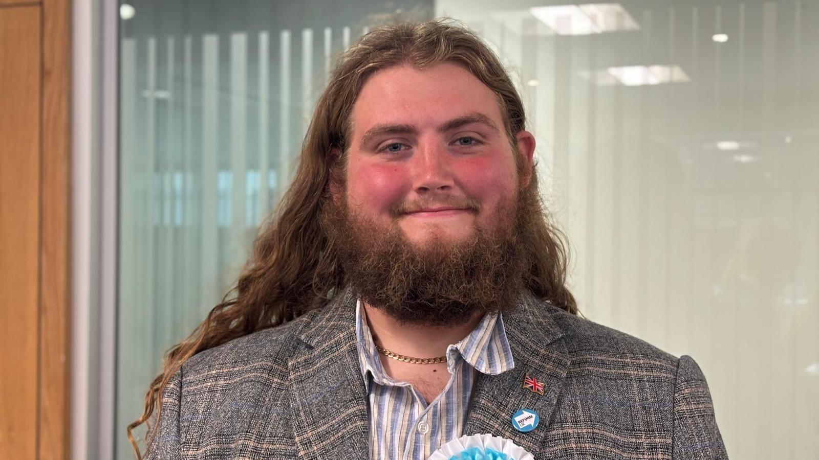 A man with rosy red cheeks smiles directly towards the camera. He has long, wavy gingery-brown hair and a large bushy beard. He is wearing a grey checked blazer, an open-collared shirt with white, blue and pink stripes and a gold chain around his neck. His lapel has a British flag pin badge, a blue Reform UK badge and the top of a white and blue Reform UK rosette can be seen at the bottom of the shot.