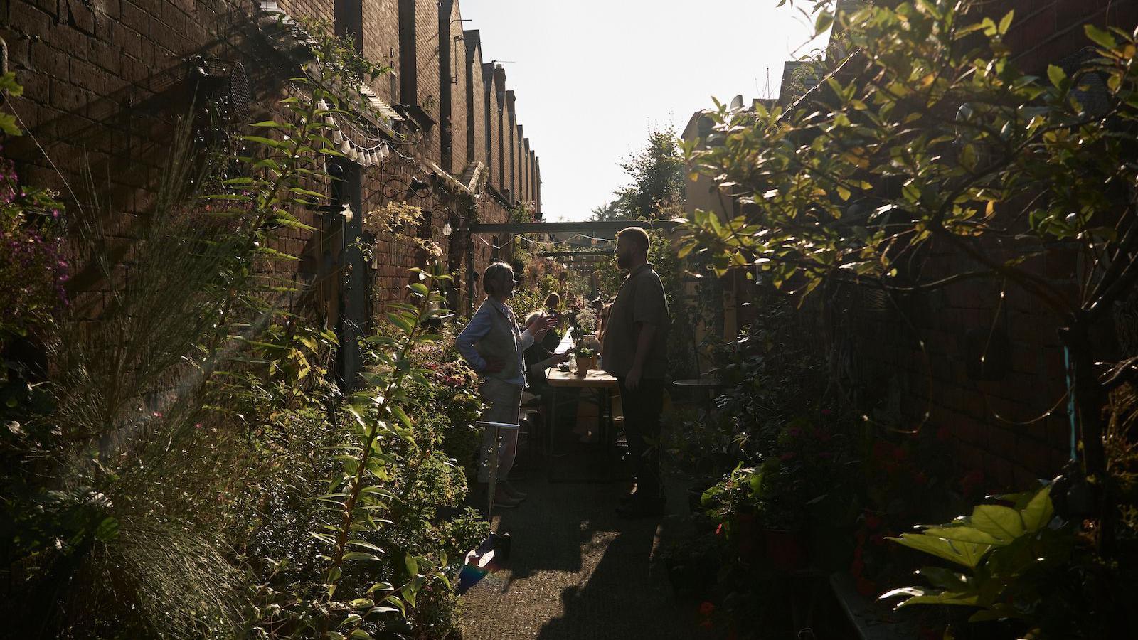 An alleyway that has been turned into a garden. People can be seen in the distance at a picnic style table. Trees and plants are along the side.