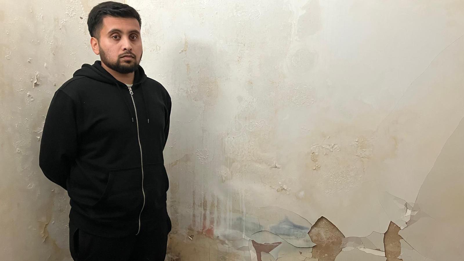 A man with a solemn expression, dressed in a black tracksuit stands in front of an internal wall. You can see paint peeling away in large chunks and blue, orange and red discolouration.