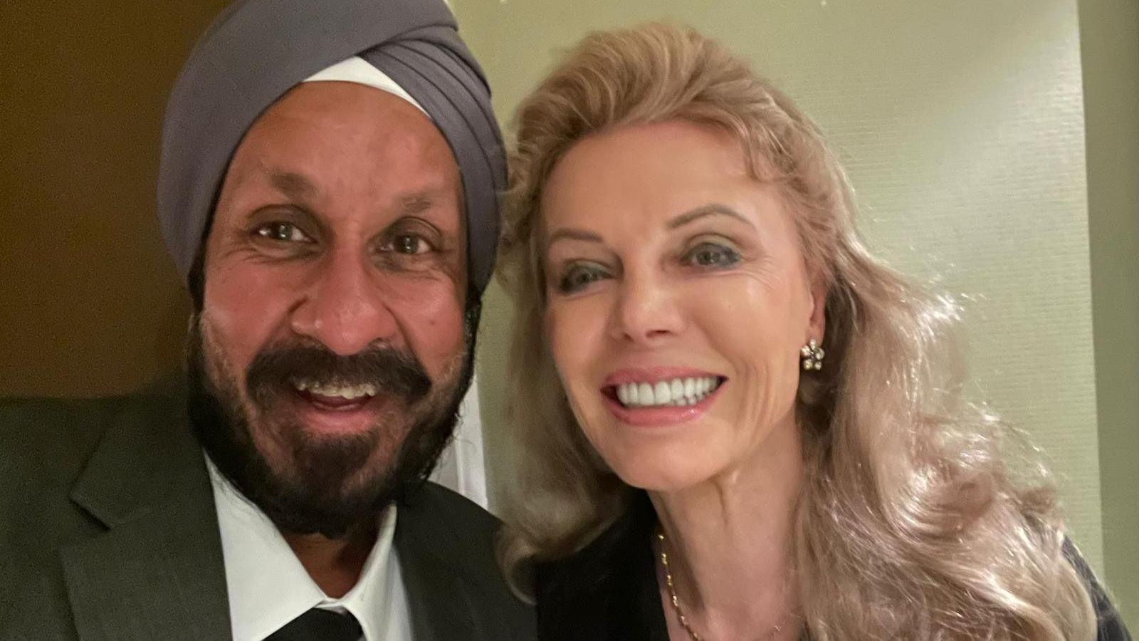 Del is taking a photo of himself and Kristina. They are both smiling at the camera. Del is wearing a light purple turban with white underneath it. He has dark facial hair, brown eyes and is wearing a suit. Kristina has long blonde hair, earrings and a gold necklace.