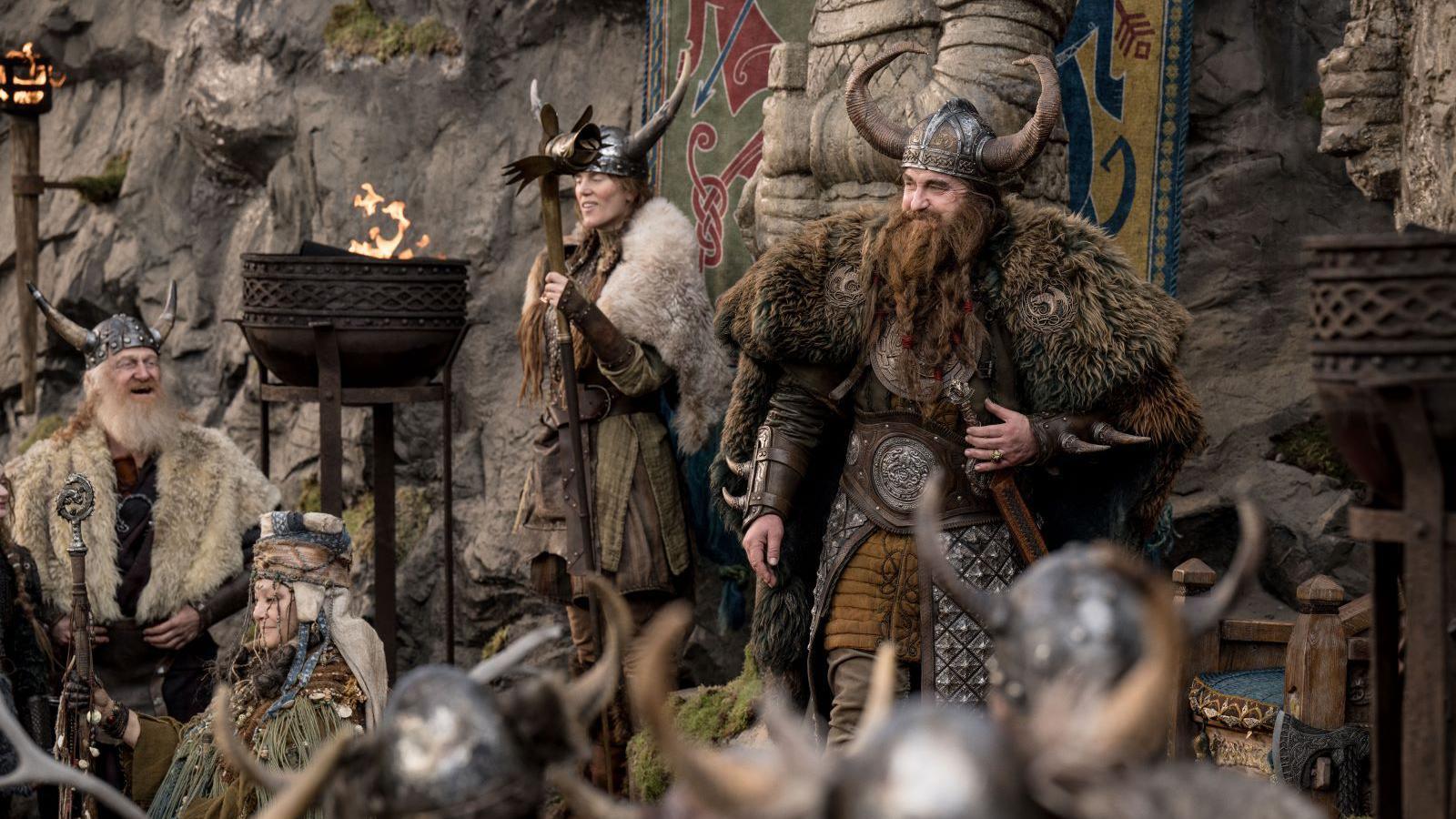 A man dressed like a viking with a horned helmet, and draped in animal skins. He has a long beard and a woman stands next to him dressed in a similar fashion. People stand around them and a bowl with fire sits in front of them.