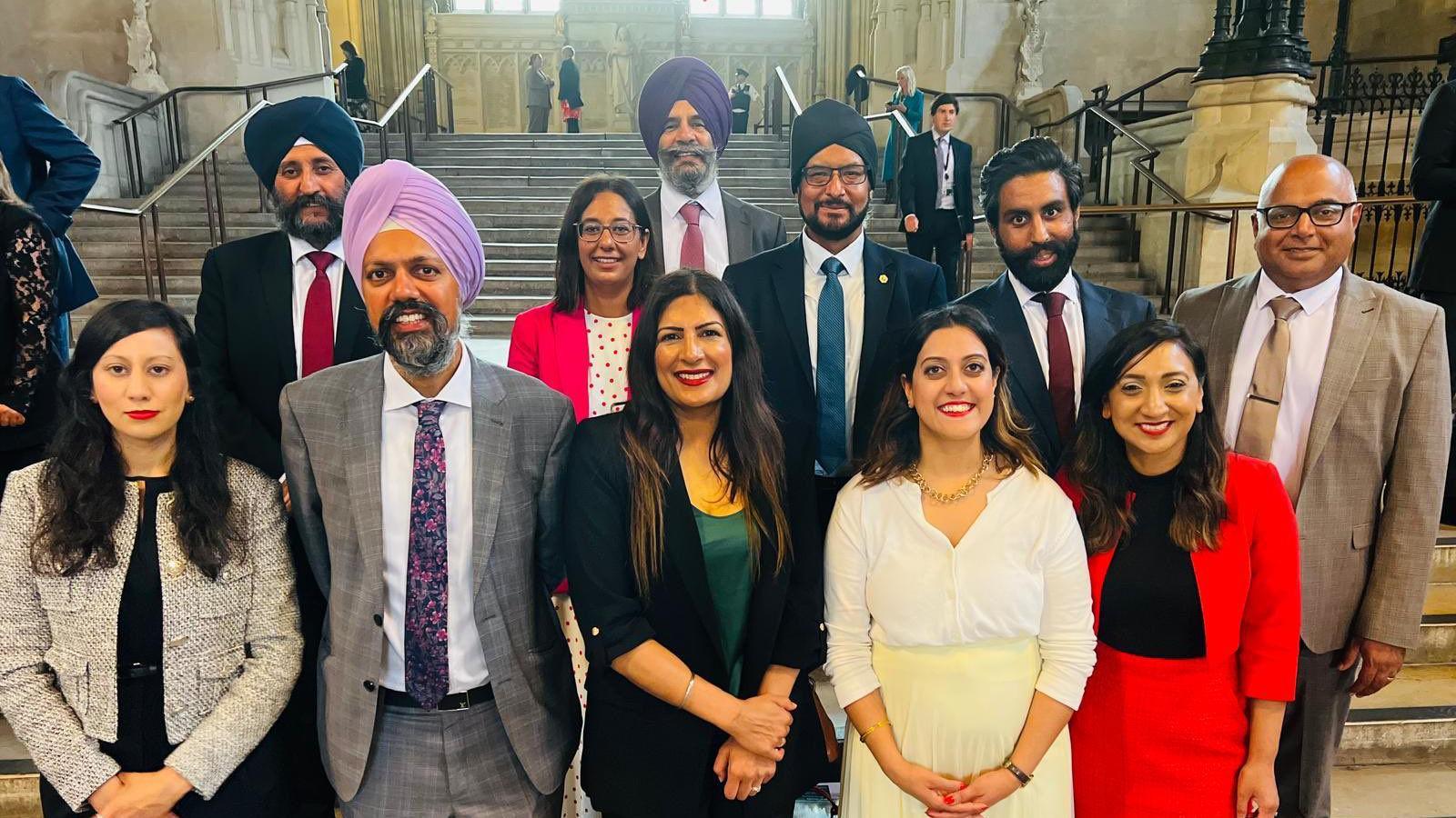 Five women and six men, four of whom are wearing turbans, pose in front of a set of steps inside the House of Commons