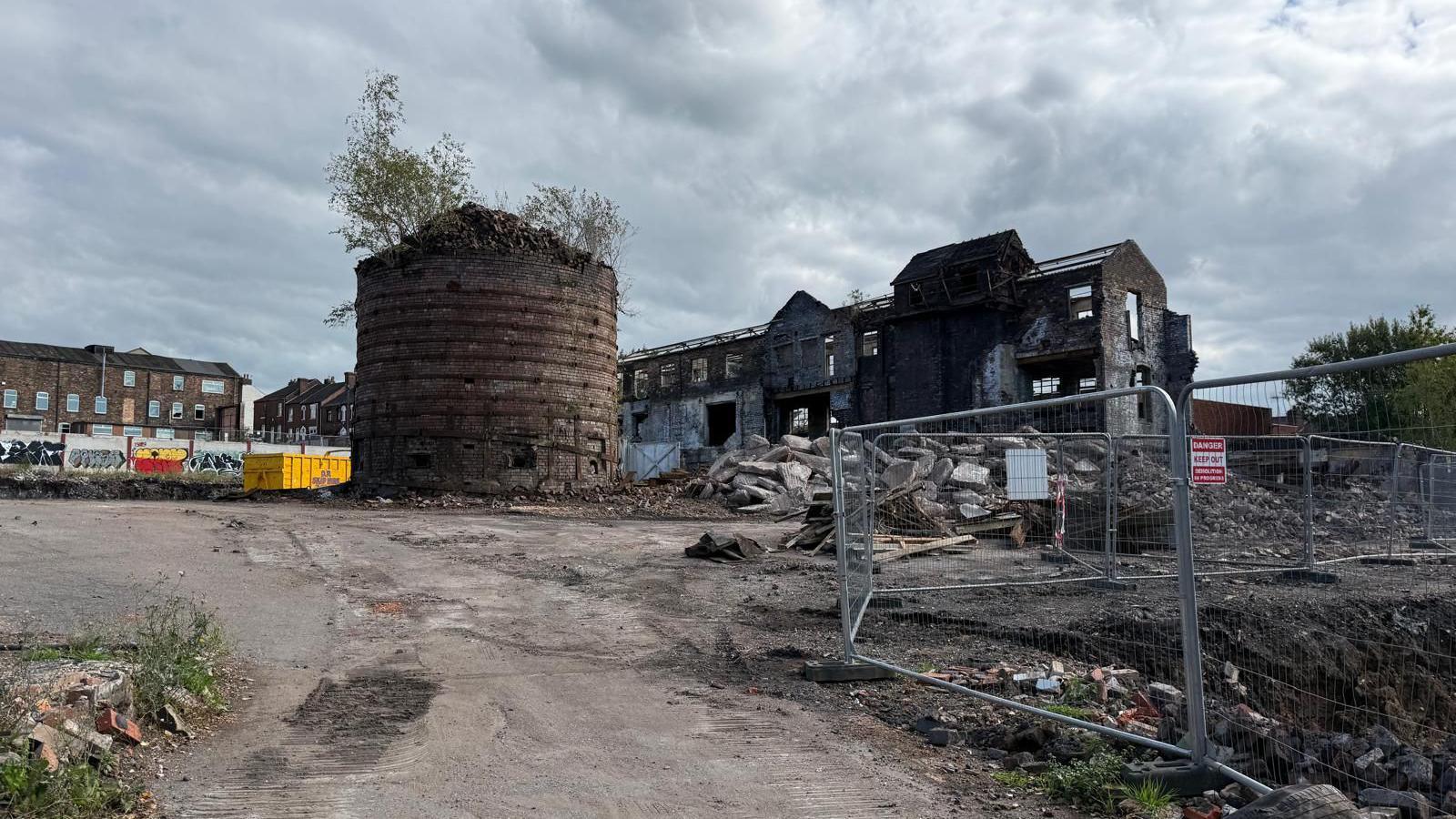 A building site, with metal fencing to the right hand side of the image and part of a bottle-shaped kiln in the middle of the site. There are derelict buildings nearby and some other buildings in the background as well as construction containers.