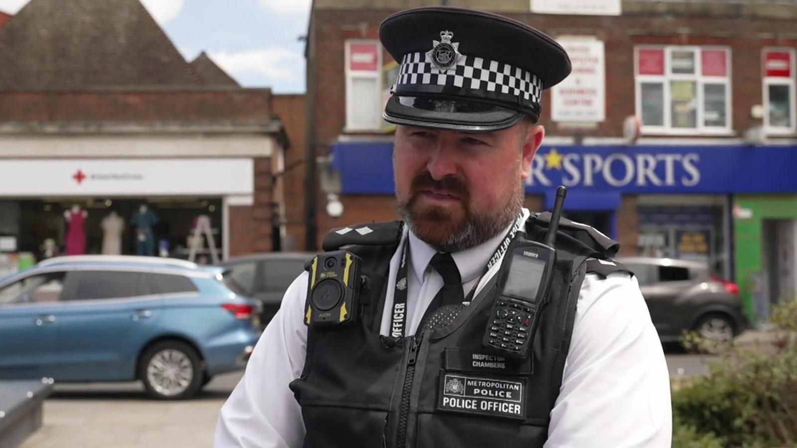 A middle aged bearded man in a police uniform being interviewed in front of a row of shops.