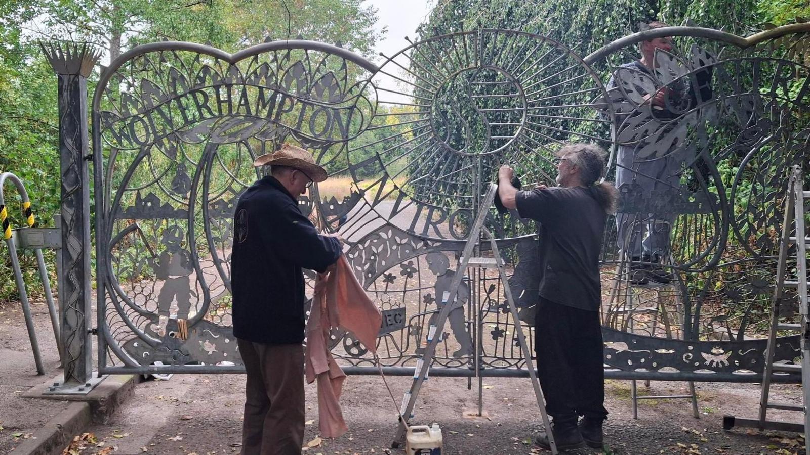 Two people seen from the back working on a metal gate. A ladder can be seen between them. The gate features ornate metal work with leaves, birds and flowers. One other person can be glimpsed on the other side of the gate carrying out work. The gate is at the entrance to a path lined with lots of trees and bushes.