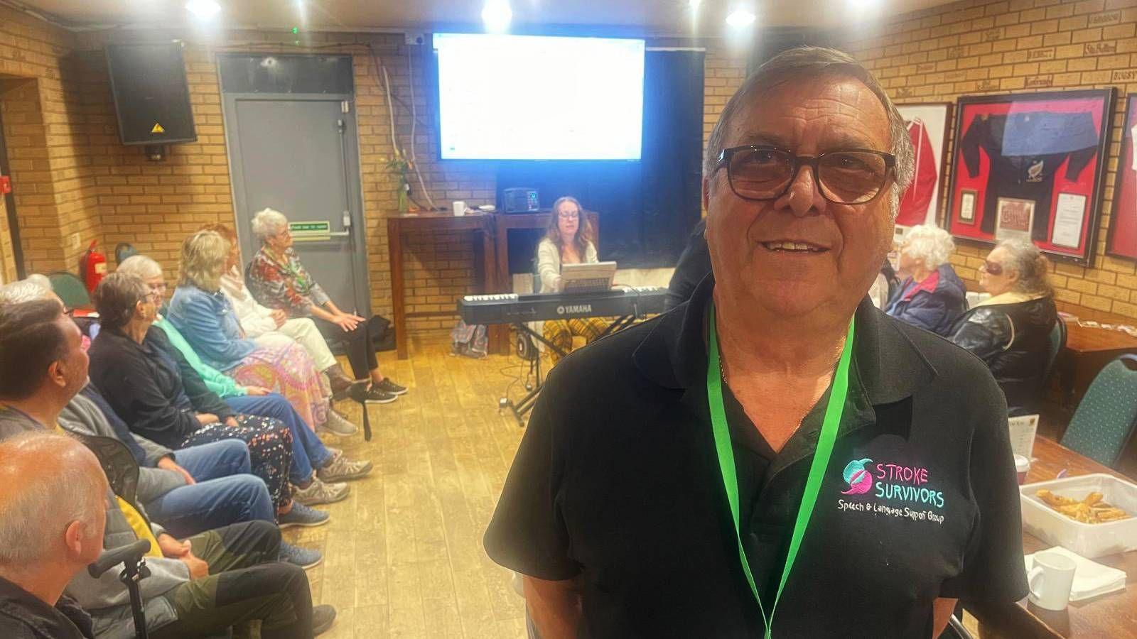 A man in glasses, a black polo and a green lanyard is standing in front of a group of people sat on chairs arranged down two sides of a small function room. There is keyboard and pianist in the centre of the room and a large television screen behind her. Framed rugby shirts and speakers adorn the walls and there is a grey fire exit door at the end of the room.