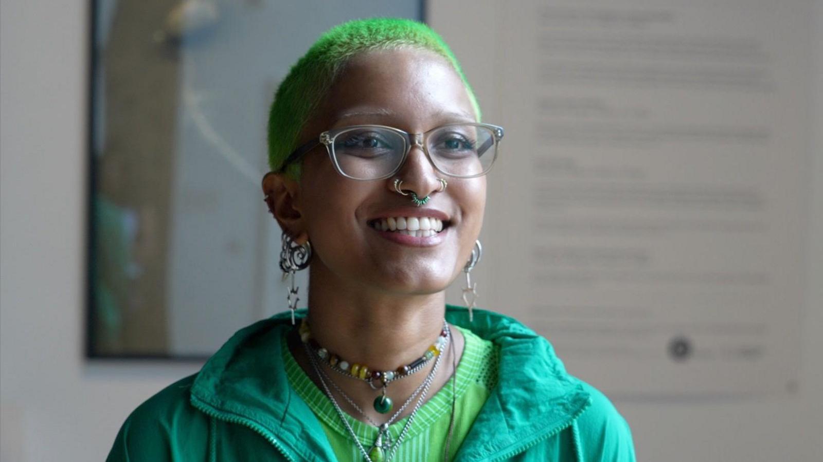 A woman with short green hair wearing glasses, a green top and green jacket. She is wearing multiple necklaces and earrings and has her septum pierced. She is smiling at the camera.