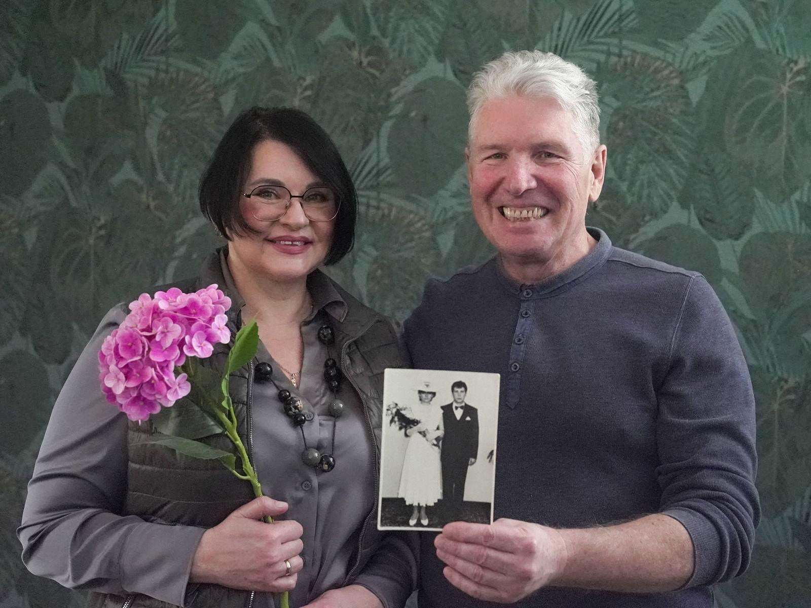 Iryna and Serhiy photographed in 2026, looking at the camera and smiling. Iryna has chin-length dark hair and glasses and is holding a pink flower. Serhiy has grey hair and is holding up a photo of them side-by-side at their wedding. 