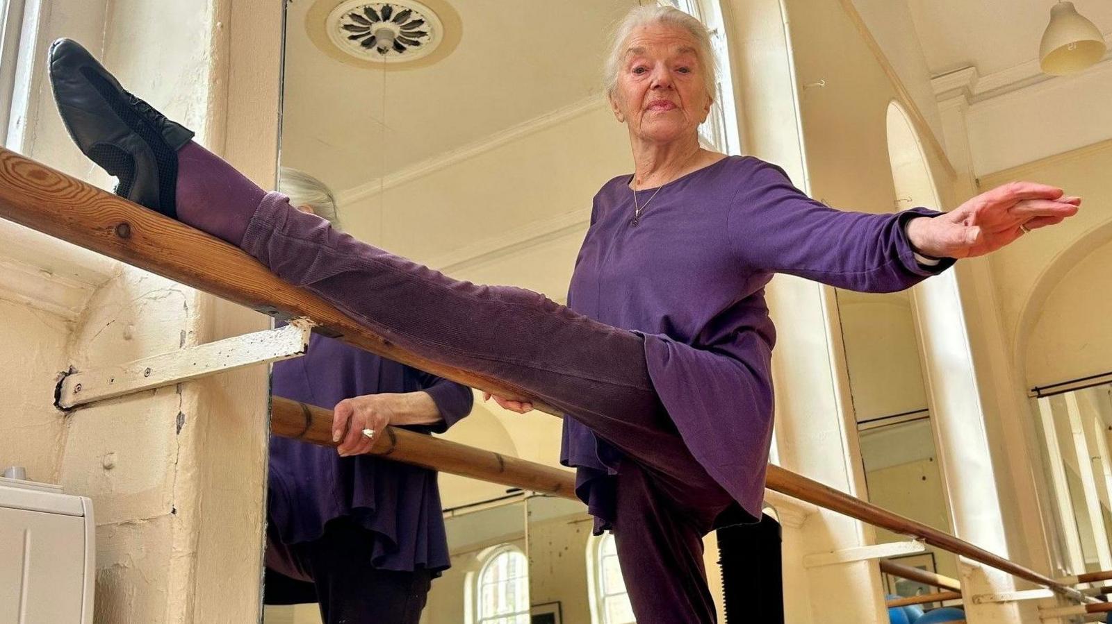 Sheena dressed in purple with her leg up in the air on a ballet barre in front of a mirror.