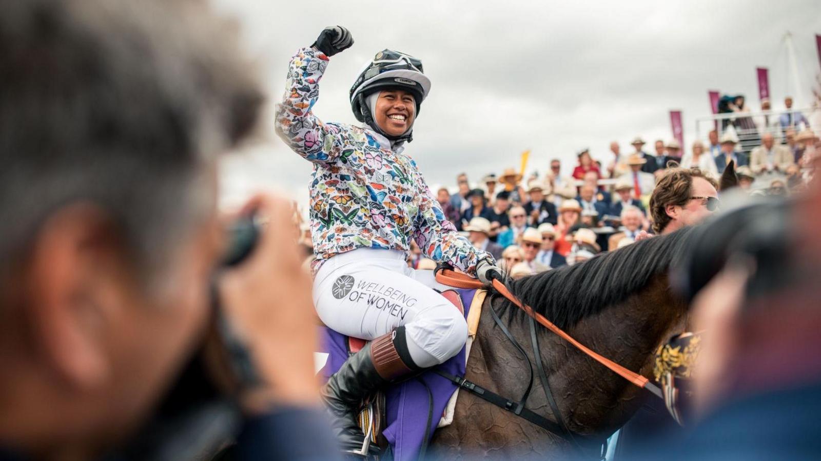 A woman wearing a hijab and a helmet and a colourful horse-riding top. She is riding a horse. There is a crowd in the background. 