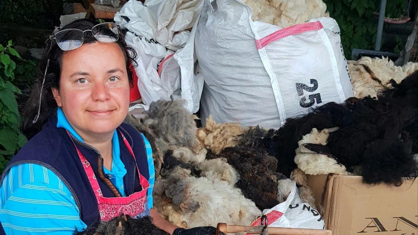 A woman in her late 30s with dark hair tied back. She has glasses on her head and is wearing a blue striped shirt with a pink apron on top. She is surrounded by bags of wool.  