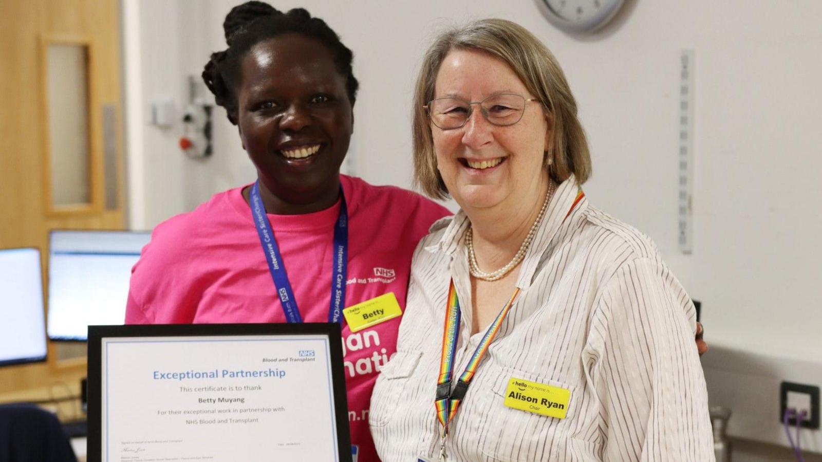 Alison Ryan with a colleague, Betty. They are stood side by side, smiling at the camera. Betty is wearing a pink organ donation t-shirt and holding a framed certificate.