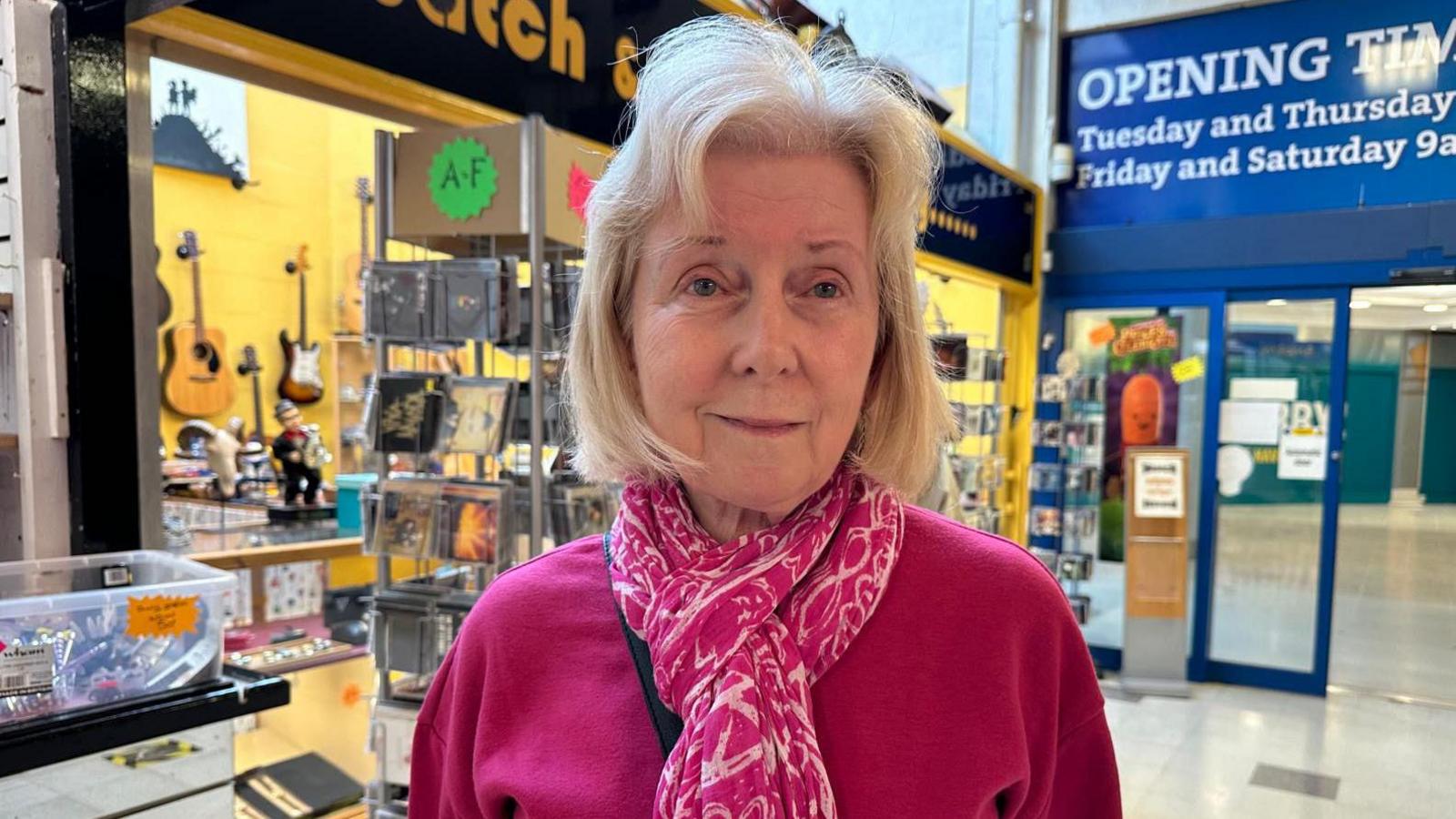 A woman with blonde hair, a pink and white scarf and a pink top stands in front of a guitar store in Stafford indoor market.