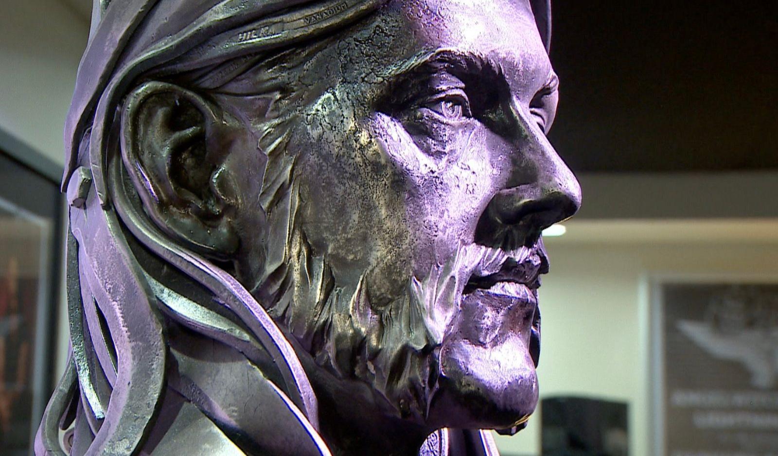 A close-up in profile of a metal bust of Lemmy from Motorhead, with purple light shining on it. The bust is silver in colour and has long hair and a beard.