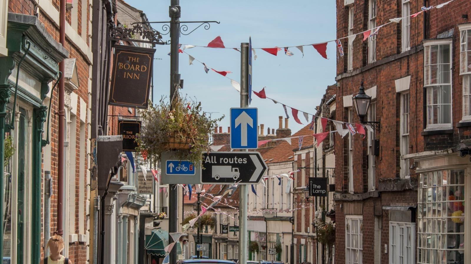 Street scene with shops and bars either side of a narrow road, with bunting strung up between the buildings. There are various road traffic signs, including one which reads: "All routes".