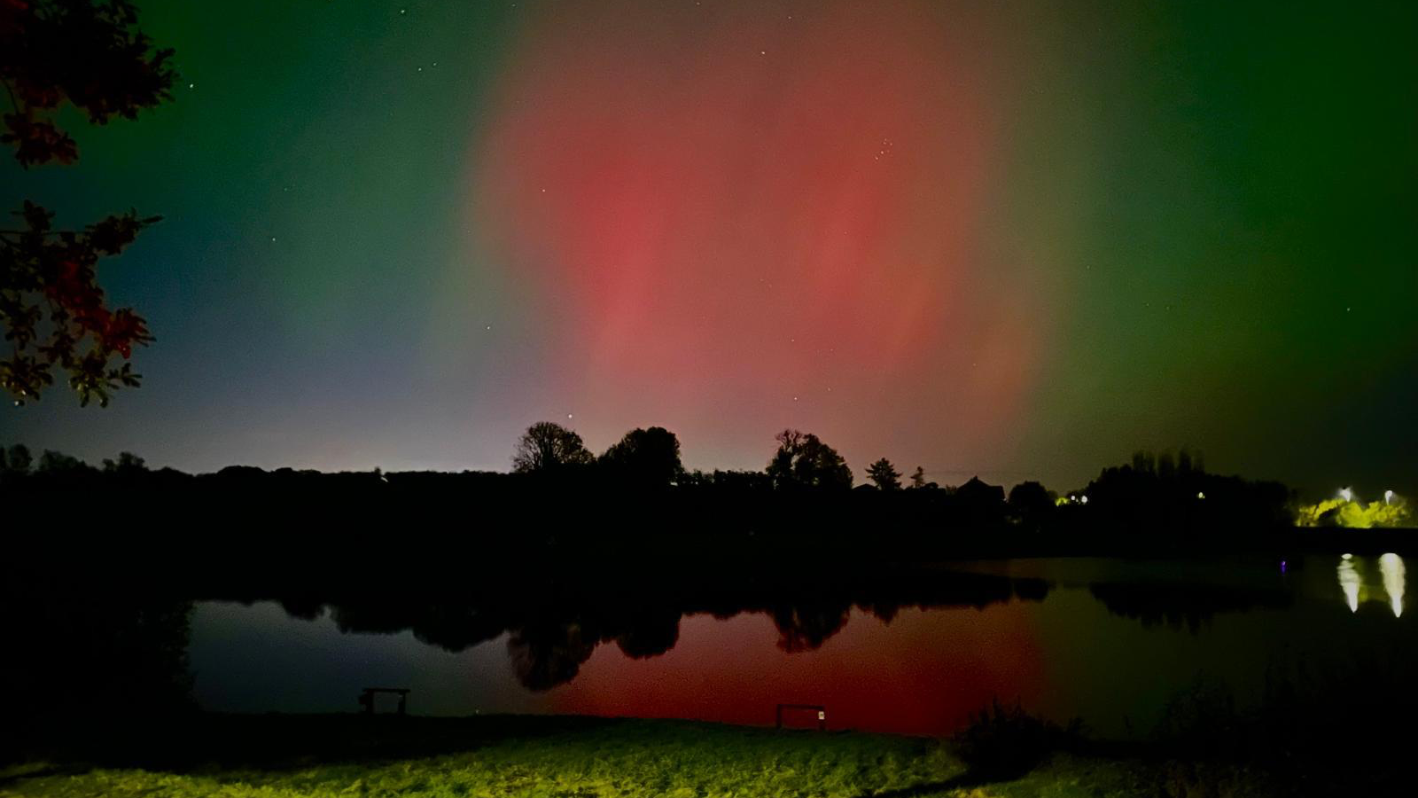 Northern Lights in the sky above a dark body of water. There are streaks of blue, green and red in the sky. 