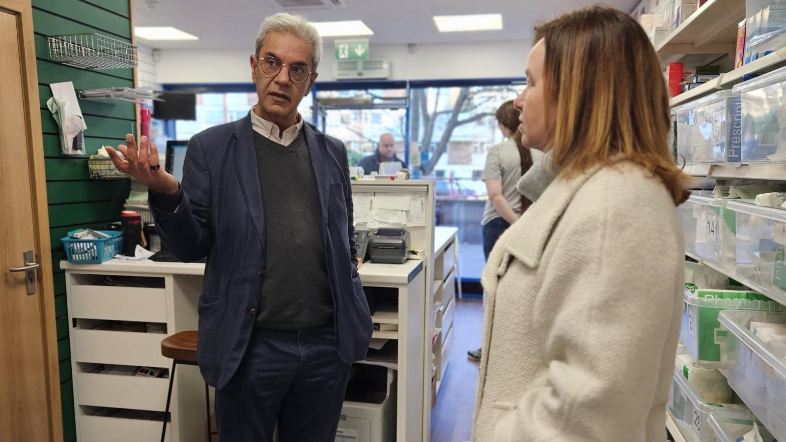 Marie Goldman and Dipak Pau inside a pharmacy in Essex. Mr Pau is pointing at something while talking to Ms Goldman.