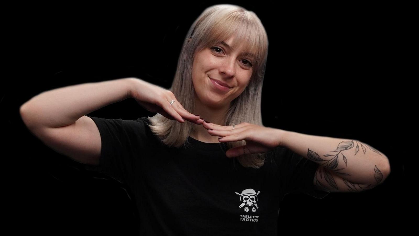 A woman with shoulder length blond hair wearing a black t-shirt poses in front of a black studio background. A white "Tabletop Tactics" logo is visible on the t-shirt. She's smiling and has the finger tips of each hands touching under her chin, her palms down and elbows up.