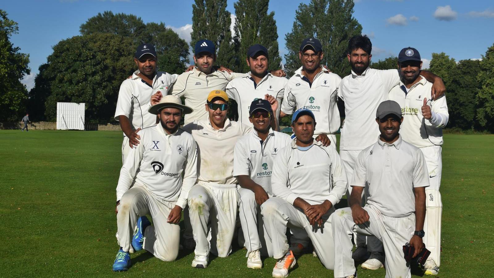 Oxenford's Saturday 1st XI team of 11 male players smiling in a field under blue skies. Some of them wear glasses. One player is giving thumbs up.