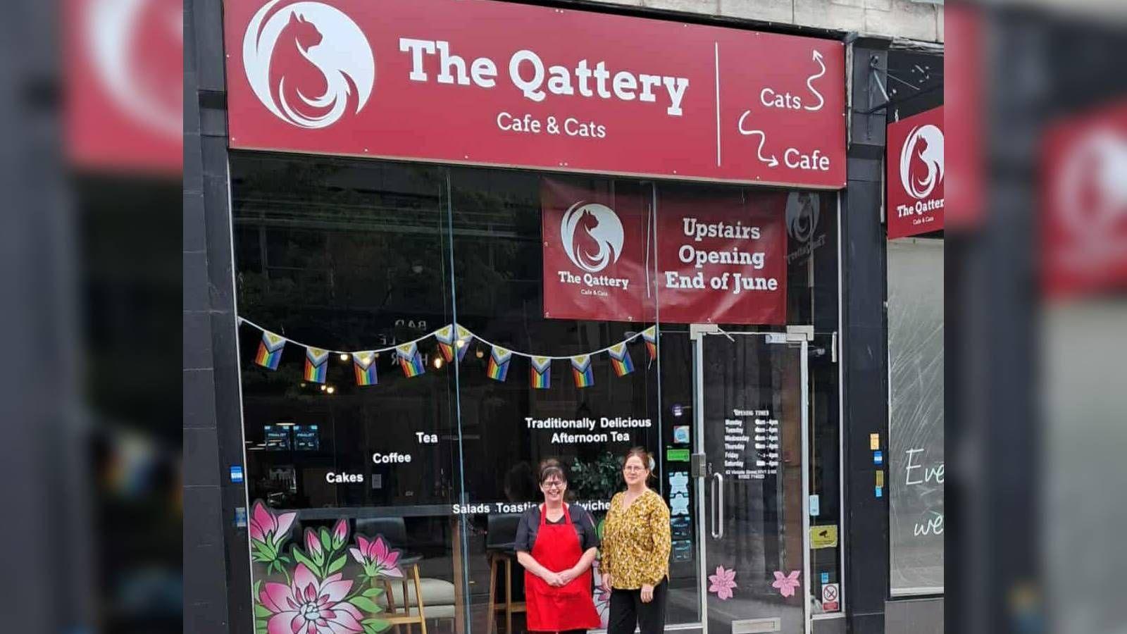 Two women stand outside of a cafe with a red sign saying The Qattery. The woman on the left wears a red apron over her shirt while the woman on the right wears a yellow shirt. Progress flags are hung up in the window.