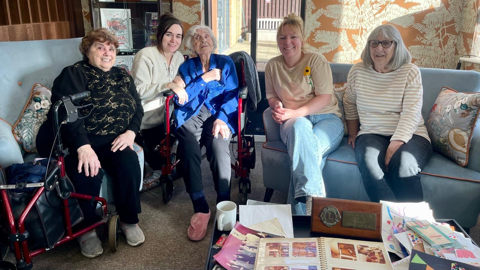 A woman with grey hair and a blue blouse is sat in a wheelchair. Either side of her are four women sat on sofas. The woman on the far left is dressed all in black with gold patterns on her jumper. Next to her is a woman in a cream cardigan and short black hair tied back. Far right is a woman in navy trousers, a yellow-and-white stripped jumper, black rimmed glasses and grey hair. Next to her is a woman in jeans, a yellow t-shirt with a sunflower design and short blonde hair tied back. In front of them all is a table filled with photographs, an award and birthday cards.