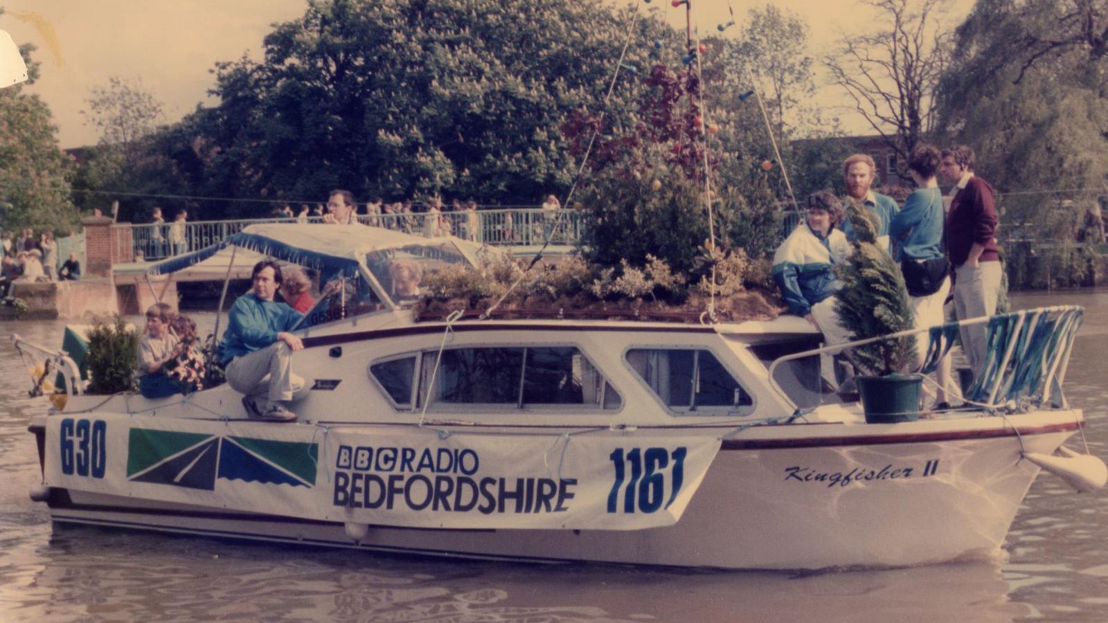 A small white boat sails along a river. It has people onboard and lots of trees as well. It has a banner advertising BBC Radio Bedfordshire.