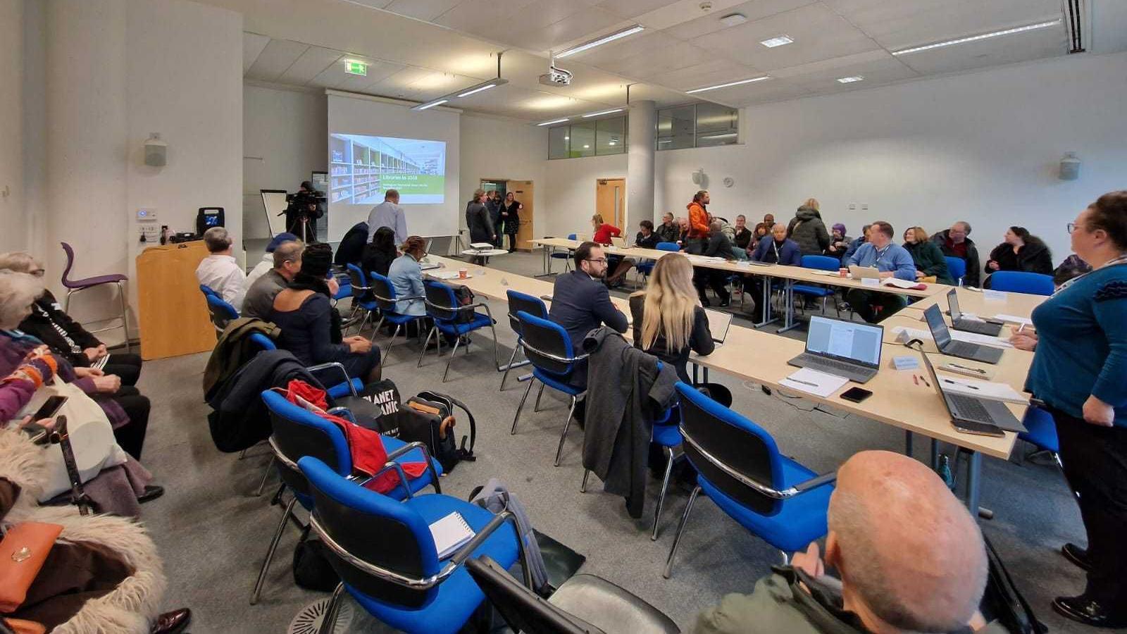 A photograph of a meeting with a large rectangular table in the middle and rows of chairs either side, full of people.