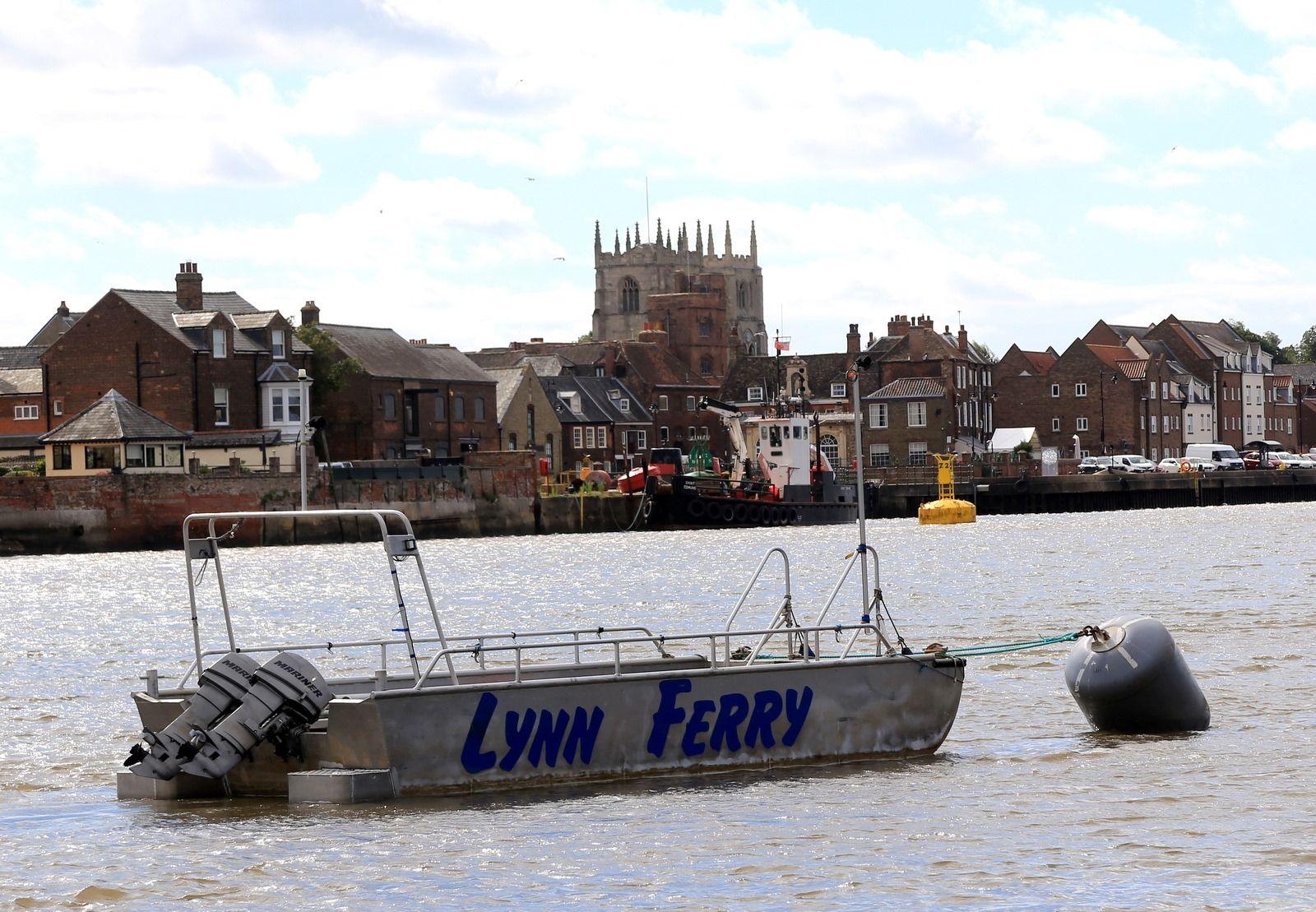 The West Lynn Ferry is a small metal boat with a outboard motors engine. It is tied to a grey buoy on the River Great Ouse. Houses and a church are on the bank behind it.