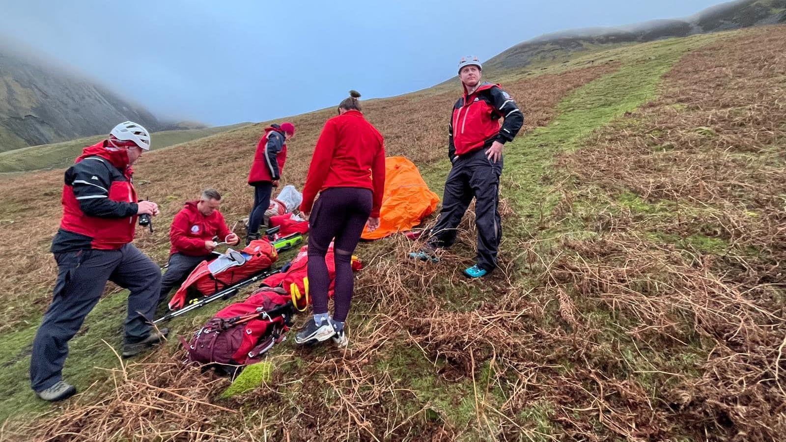 People on side of mountain is red rescue gear. They are surrounding a stretcher on the floor. 