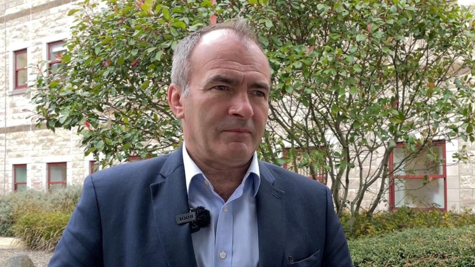 A politician with short hair in a navy suit and light blue shirt, standing in a hospital courtyard with bushes and a small tree in the background. 