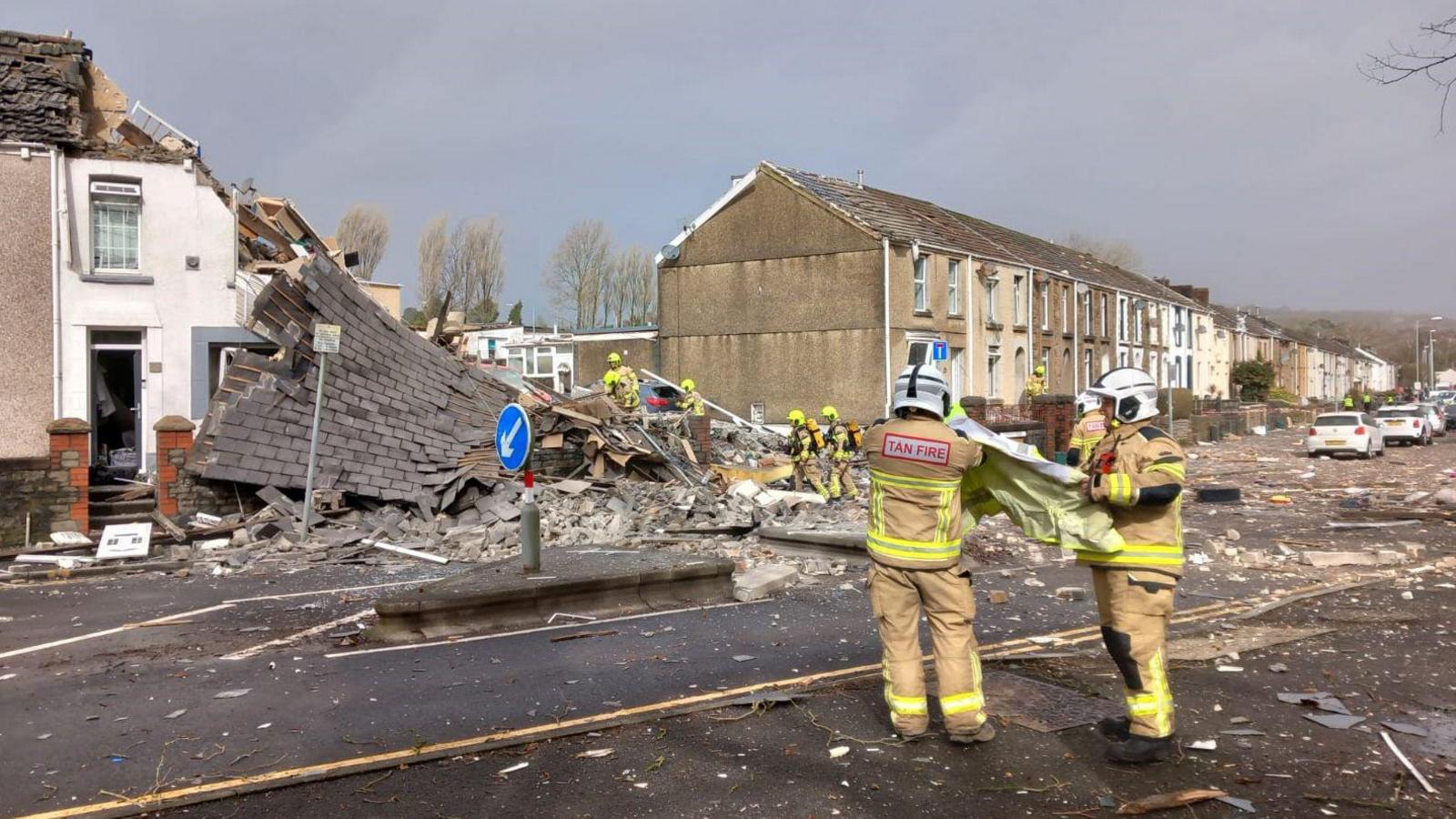 A scene showing the destroyed house, which has been flattened alongside a row of houses. In the foreground, two people wearing fire service uniforms are seen