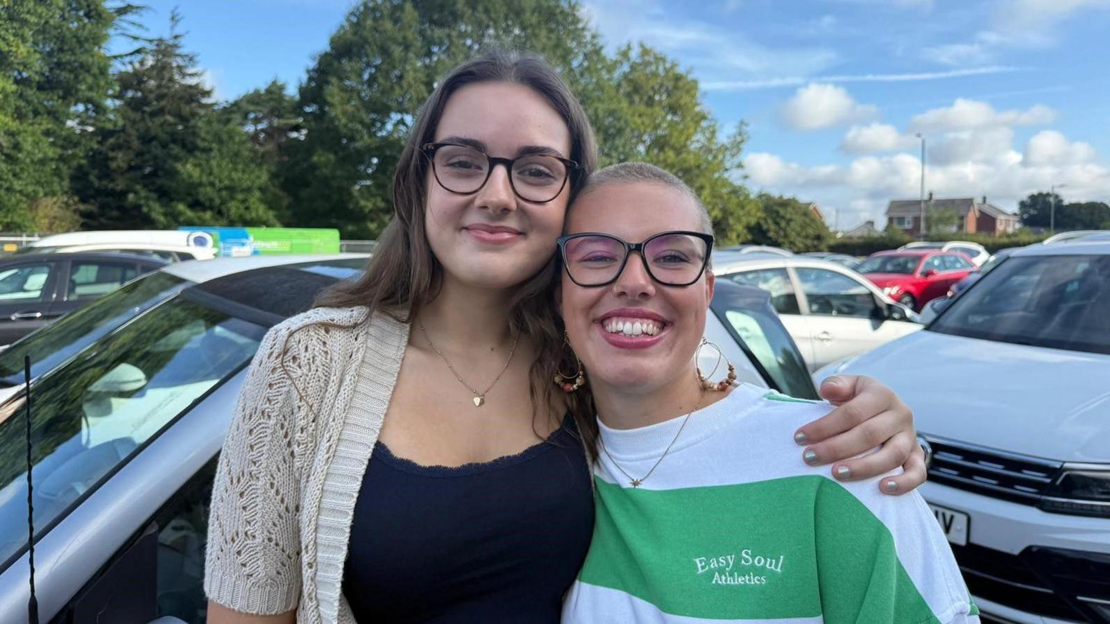 Orelia and Faith stand in the college car park. They are both smiling at the camera. Orelia stands on the left, she has her arm around Faith's shoulders. Orelia has long brown hair styled with a centre parting. She wears round, tortoiseshell framed glasses and a fine gold chain necklace, with a black scalloped edge tank top and a beige crocket cardigan over the top. Faith has shaved blond hair and wears large black framed glasses and large hooped earrings. She wears a long sleeved top which is white with a pale green stripe along the front and a logo which reads 'Easy Soul Athletics' embroidered on the chest. Behind them are a number of parked cars and some trees in the distance.