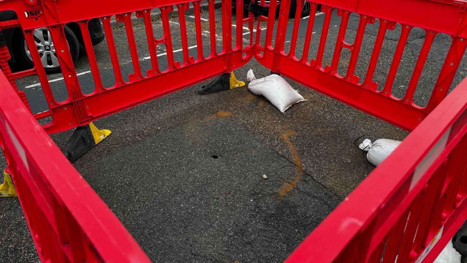 A picture of a tiny hole in tarmac in a road with red crash barriers around it, with two sandbags holding one of the four down.