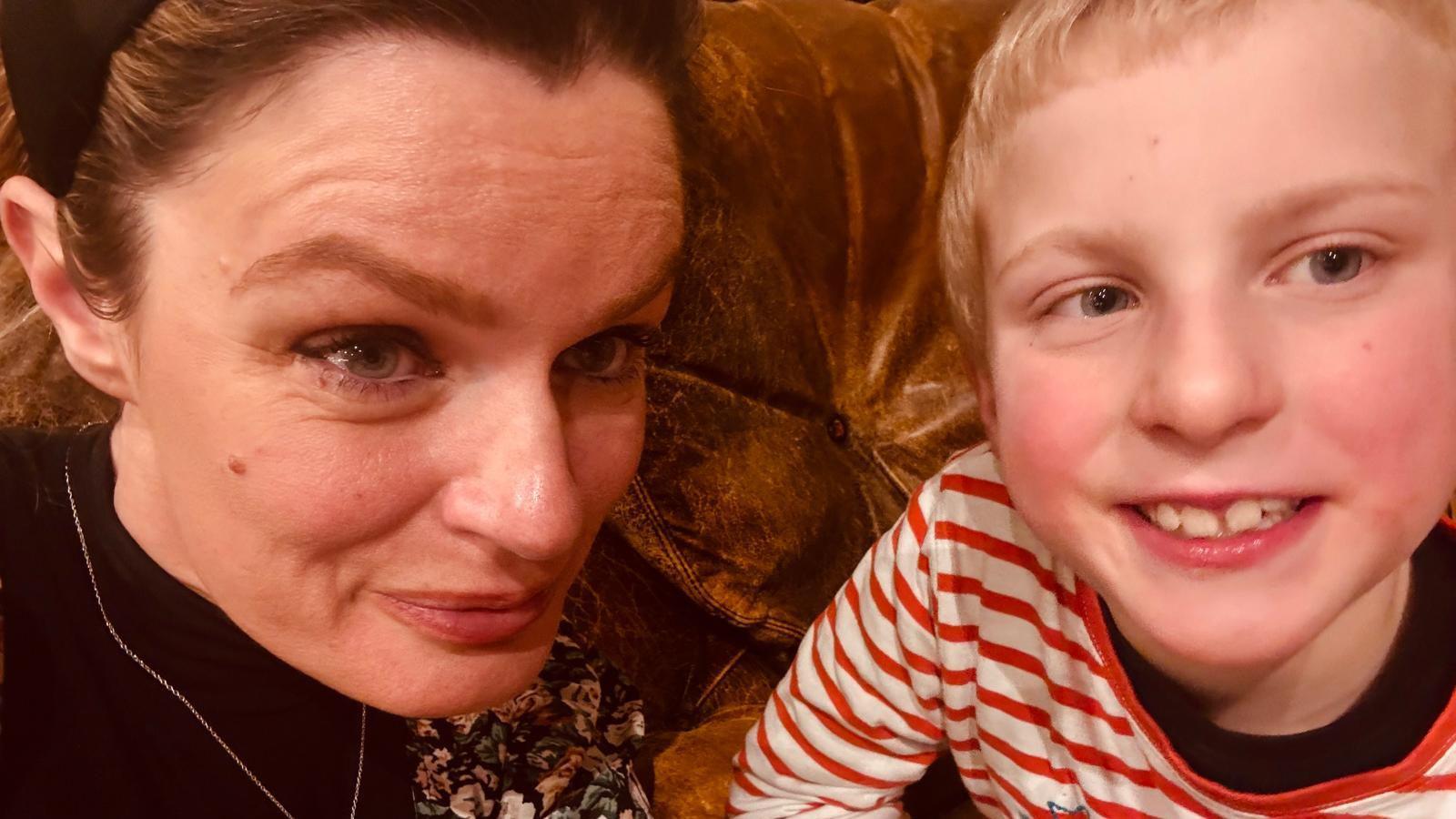 Tracy Harris, who has her blonde hair tied back and is wearing a black headband, sitting on a sofa with her her eight-year-old son Hartley who has blonde hair and is wearing a red and white striped top