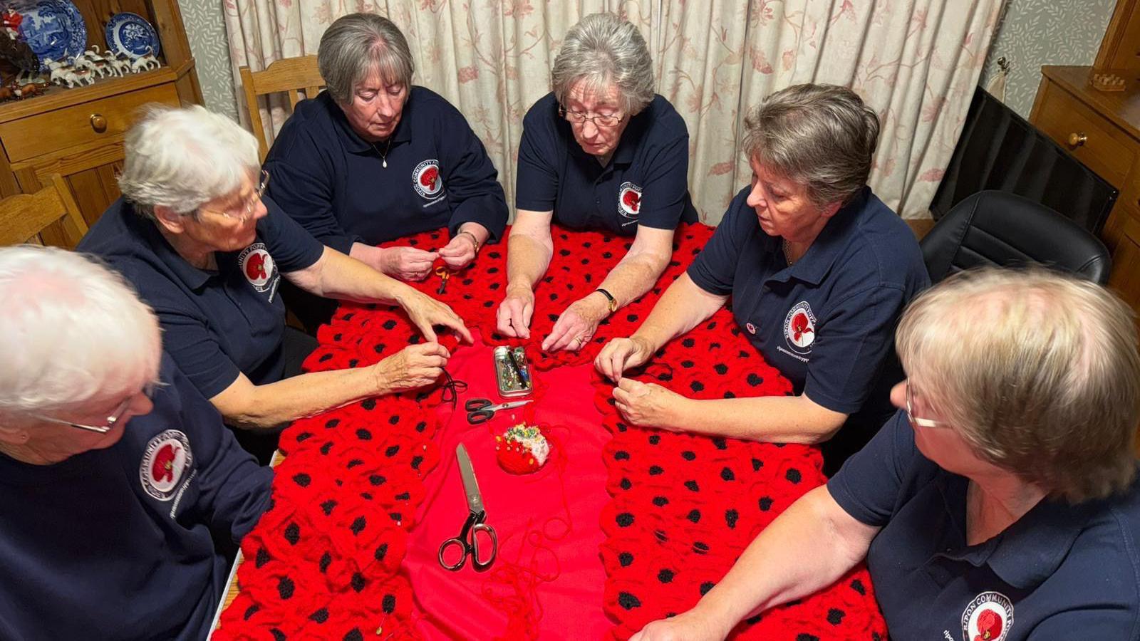 A group of six older women sit at a dining room table sewing poppies onto a garment.