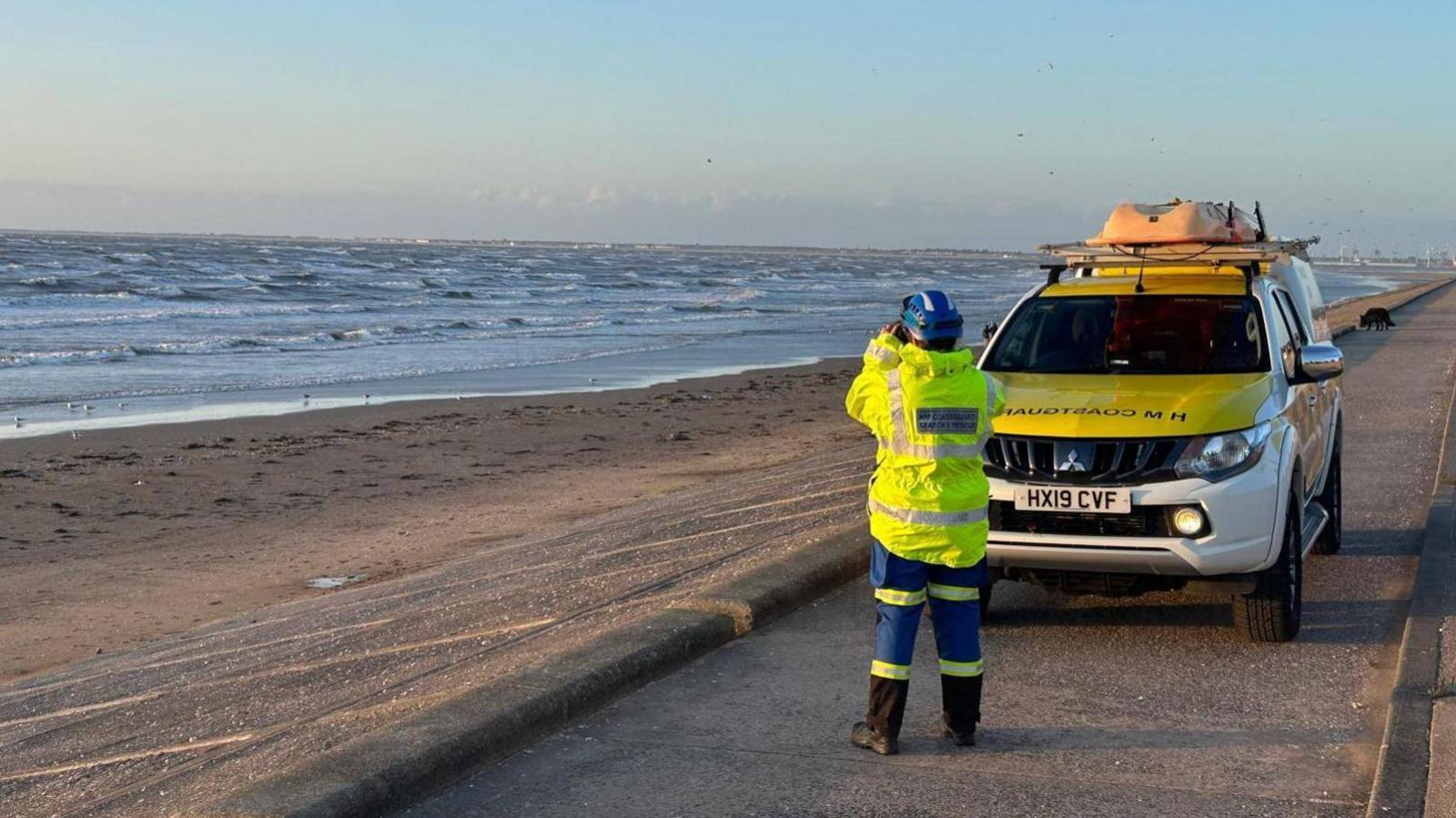 A HM Rescue team officer looking out to sea through binoculars, dressed in high visibility clothing and stood next to a rescue vehicle.