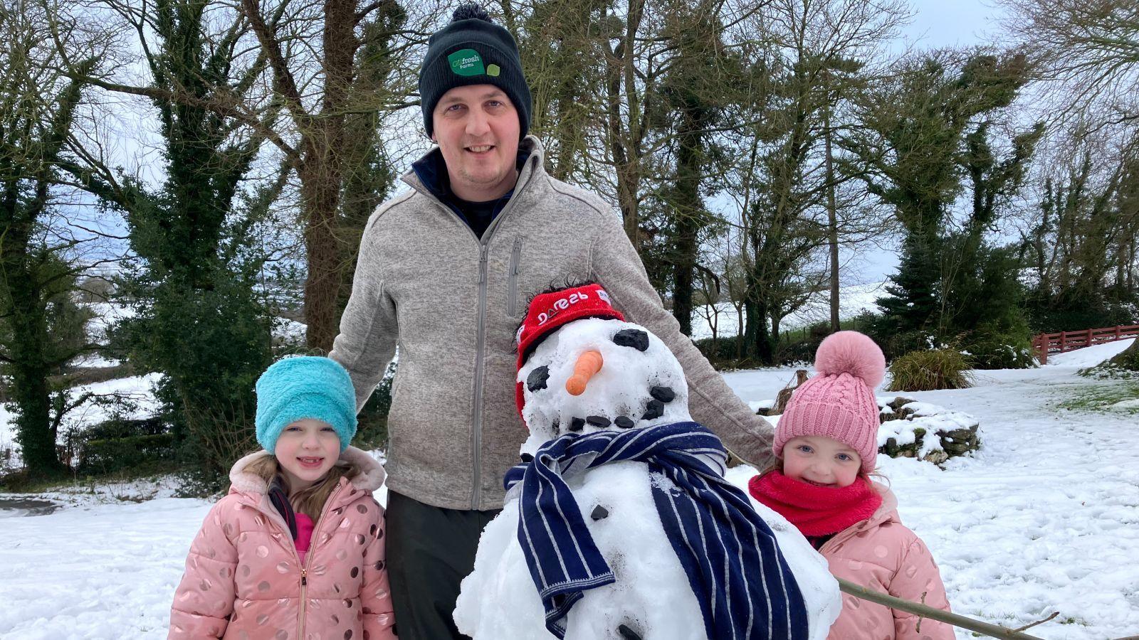 A man in a navy hat with a green crest and grey zip up top stands with two little girls in pink coats and a snow man in front of them