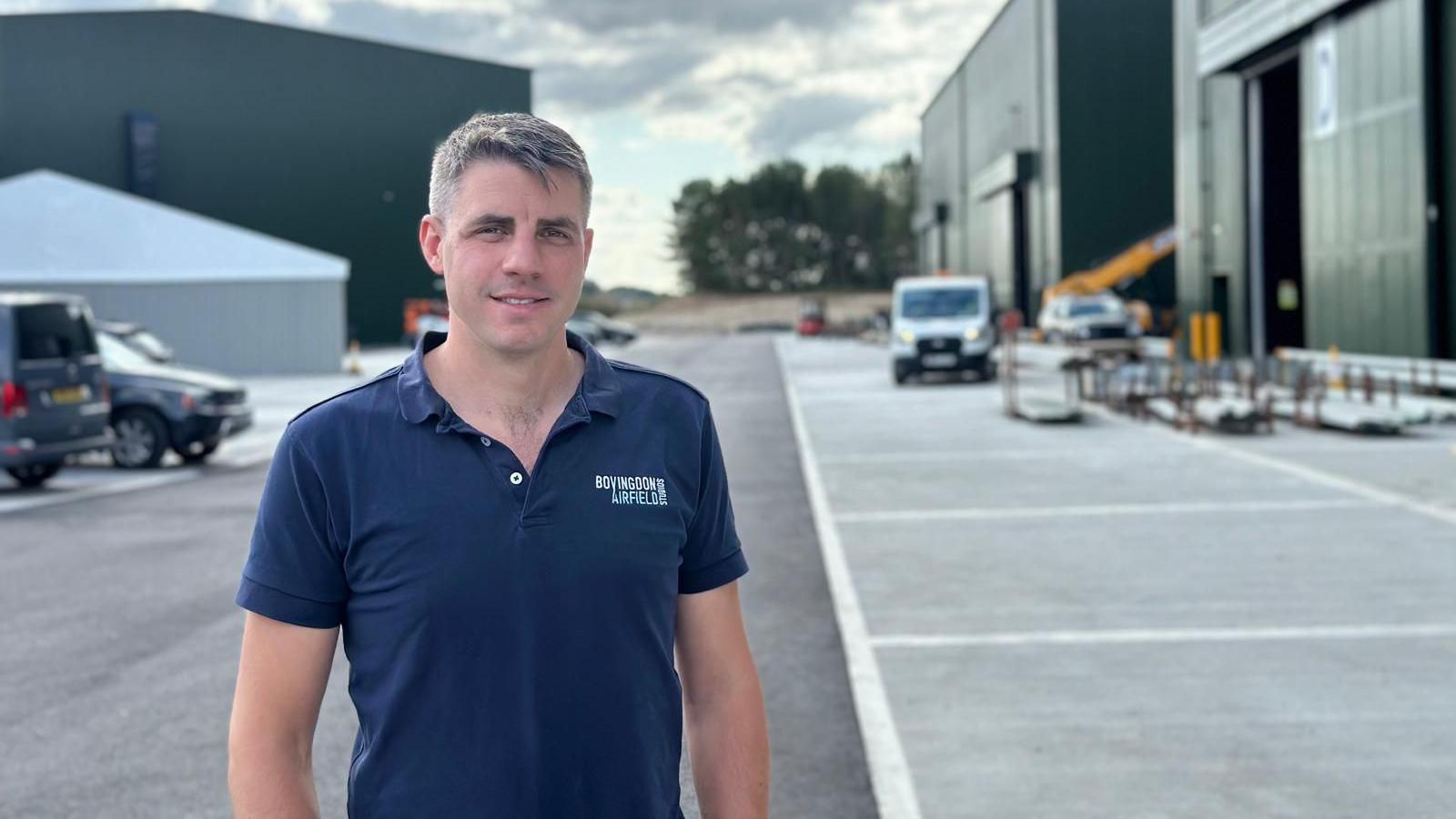 Harvey Mash has short grey hair and is wearing a navy polo shirt with the studio logo on the left breast. He's standing with large film studios behind him