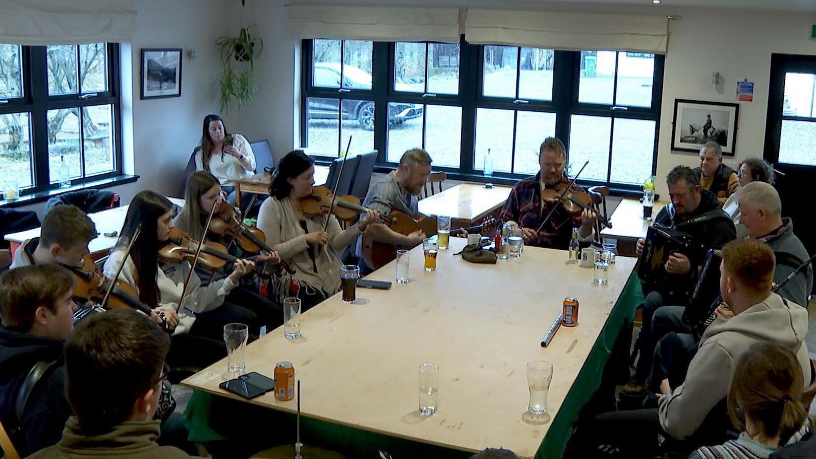 A group of musicians playing fiddles and accordions sit around a table inside the inn. There are drinks on the table.