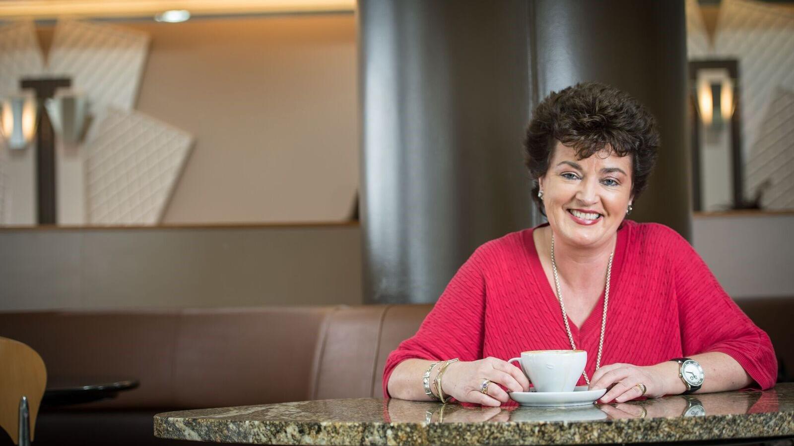 A woman with brown hair wearing a red top sits at a table, smiling at the camera. She is holding a white cup and saucer. 